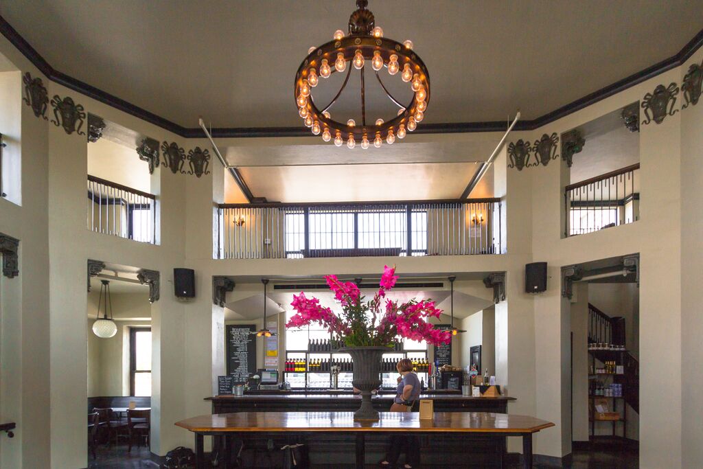A person stands at the counter of a winery in a two-story room with a Edison-style lightbulb chandelier. Bottles of Missouri made wine sit on shelves lining the windows behind the counter.