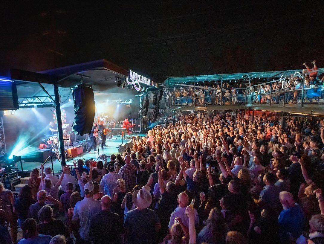 A crowd of people have their hands raised under a black, night sky. There are two levels- some people are on the ground floor and some are on a top deck. There is a stage with a band, and the top of the stage has a sign that says "Knuckleheads Saloon."