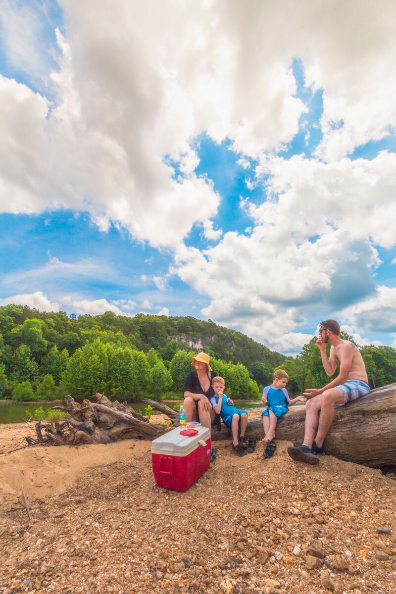 A family sits on the gravel bank of a river eating a picnic lunch. They're in swimming attire and the sky is blue. Greenery and lush trees line the opposite river bank.