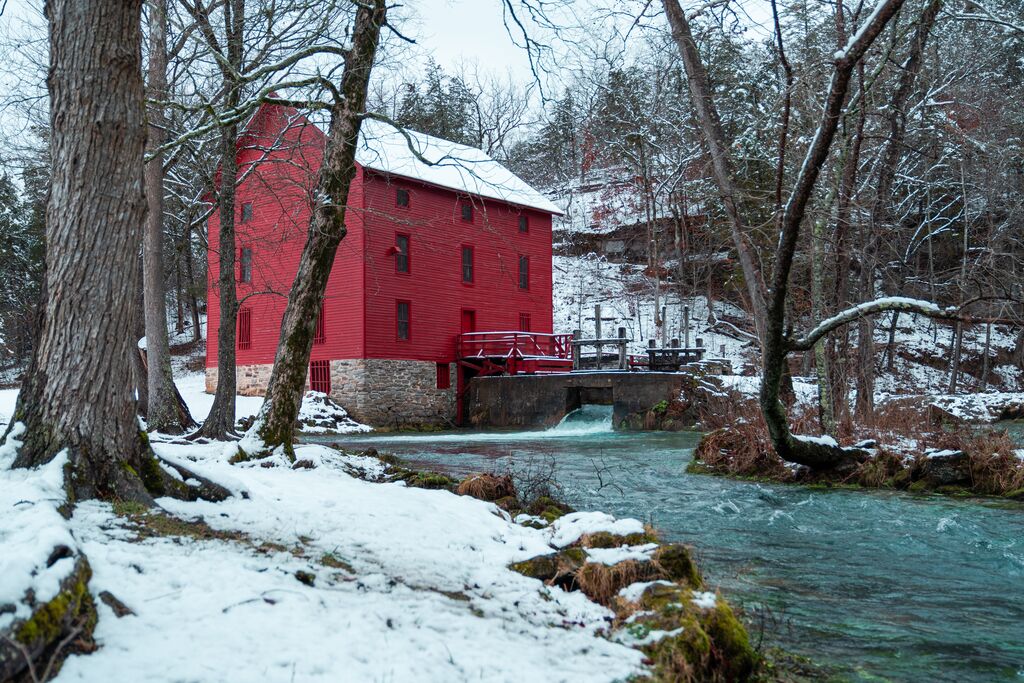 Image shows Alley Spring and Mill in Eminence, featuring a bright red mill and snow on the ground beside a body of water. 