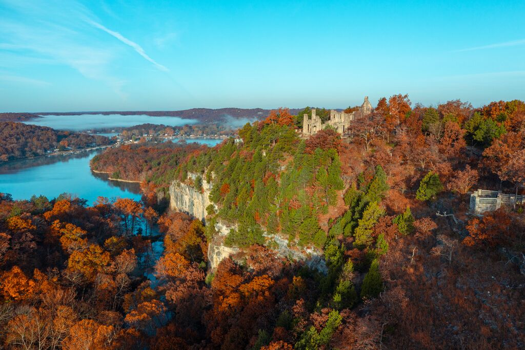 A stone castle sits on a bluff surrounded by fall leaves and a lake below.