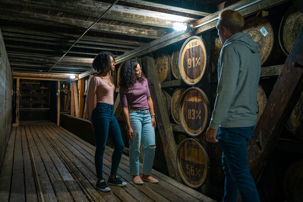Two guests stand in a long, wooden hall. Barrels lay on their sides and line the walls from top to bottom. The guests are looking at the barrels as the tour guide talks.