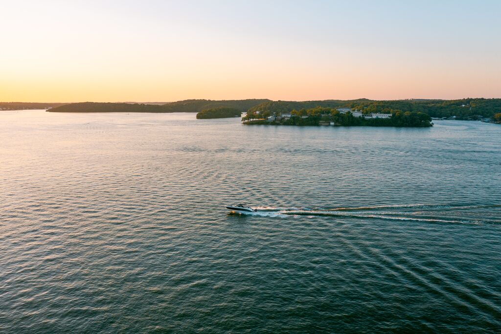 A boat cruises across the water at Lake of the Ozarks. Green, lush trees ling the banks in the background. The sun is setting, creating a yellow and pink afterglow.