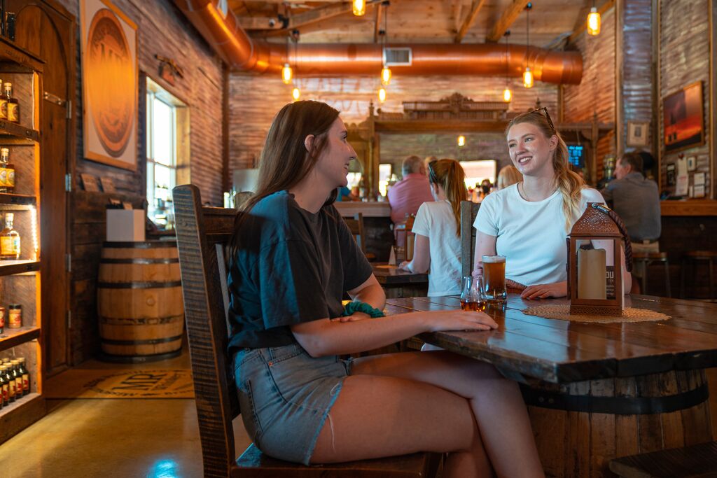 Two people sit at a wooden table. They are smiling as they sip beer and bourbon. The background is a distillery with soft, yellow lighting and a copper pipe running against the ceiling. Barrels sit along the wall.