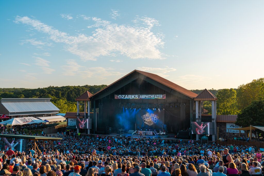A large crowd sits and watches a band perform at the Ozarks Amphitheater. The sun is shining in a blue sky filled with clouds. Green trees surround the amphitheater and food vendors line the sidewalks.