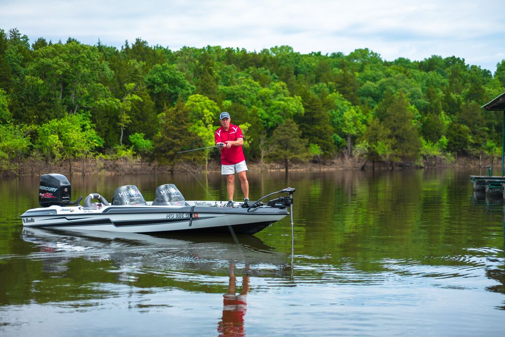There are clouds in the blue sky and lush green trees line the bank. The water is calm. A fisherman with a red shirt is standing on the edge of his motorboat, casting his line into the water.