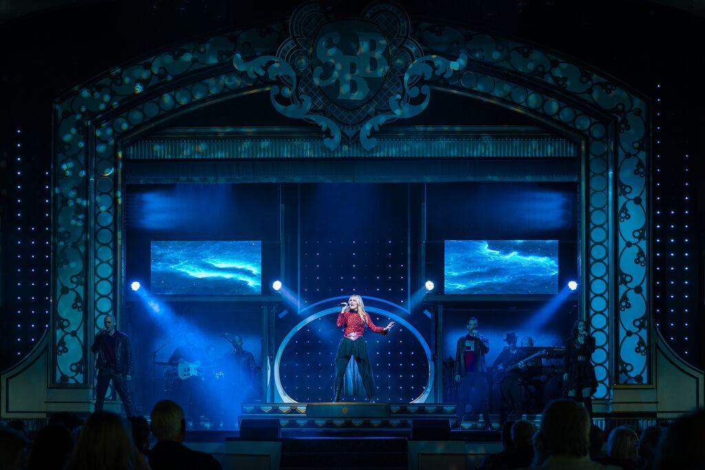 A singer with long, blonde hair and red shirt is standing in the middle of a stage. She is singing into a microphone and blue lights surround and spotlight her.