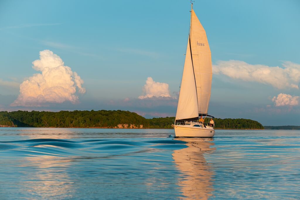 The sky is blue and has large white clouds throughout it. In the background, there is shoreline that has many large, green trees. There is a couple on a sailboat sailing, and the boat has waves beside it from the current.