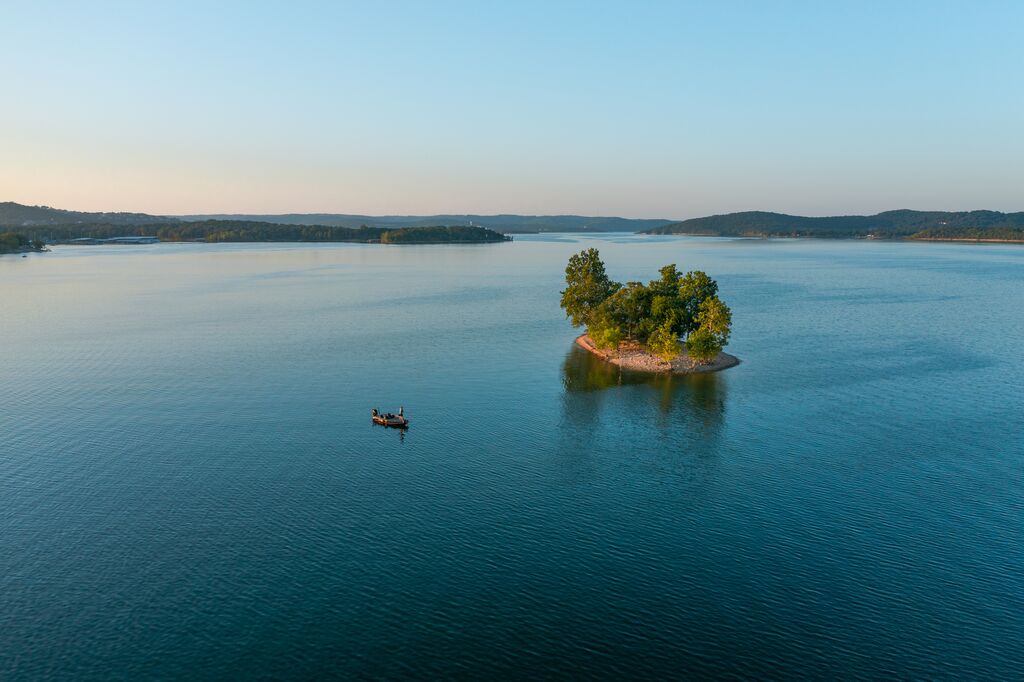 An aerial view overlooks the lake. In the distance are rolling hills with dark green trees. The sky is light blue and the sun is beginning to set. In the middle, there is a little island with green trees on it and a bank. There is a person standing in their boat not too far from the island.