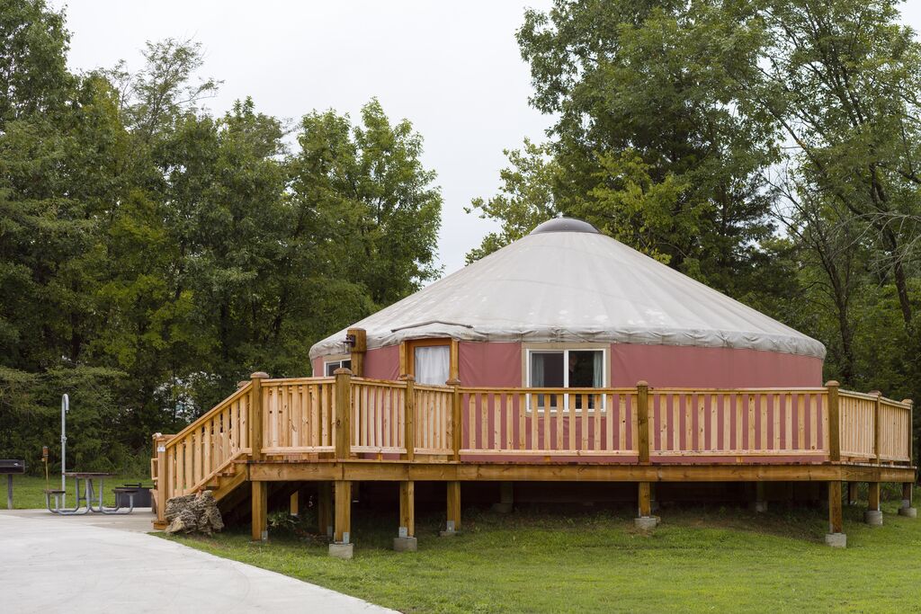 A pink yurt is surrounded by green, lush trees. The yurt has a wrap-around porch that leads to a paved parking spot.