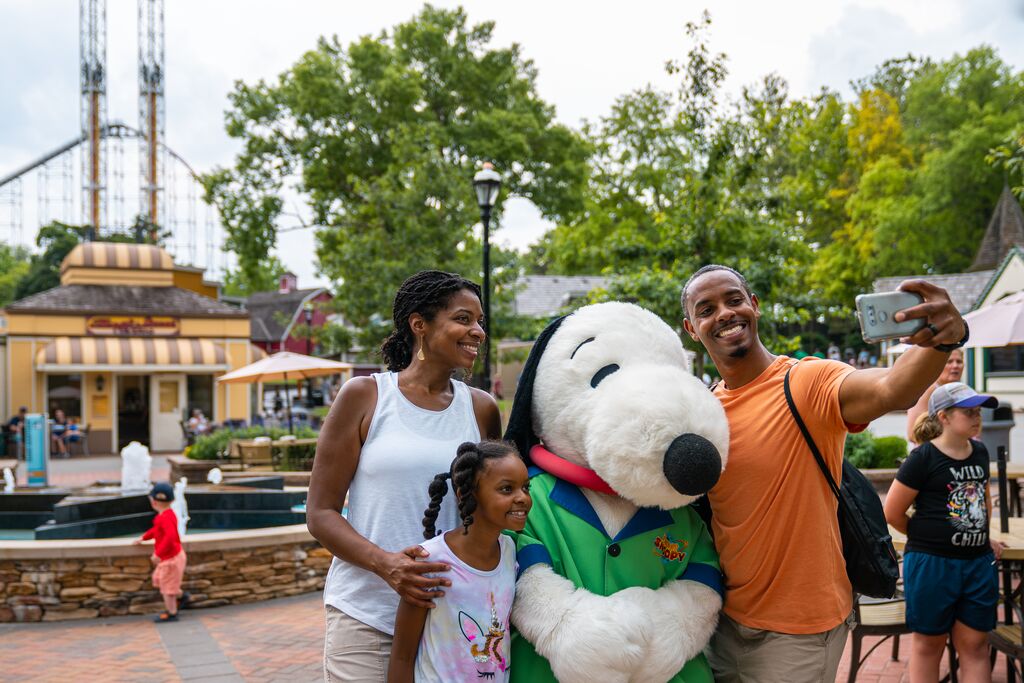 Family poses for a photo with Snoopy with food shops and amusement rides in the background.