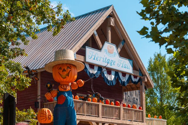 A pumpkin scarecrow waves at guests with a building covered in pumpkins in the background. 