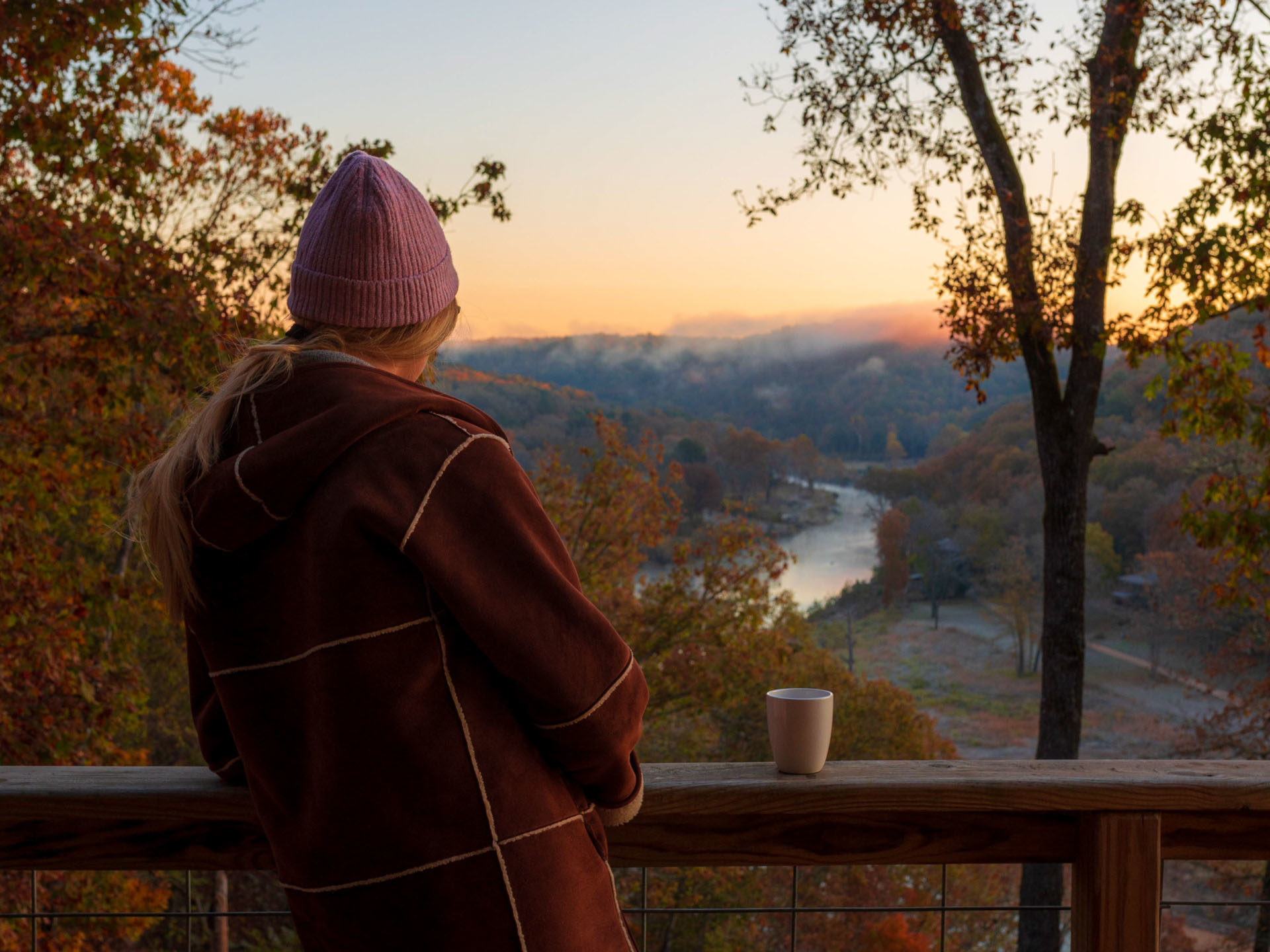 A woman with long blonde hair wearing a beanie and heavy coat stands with a mug overlooking a river valley and rolling hills at sunset. The leaves have changed color for fall.